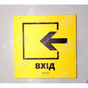 Тактильна табличка жовтого кольору з чорною стрілкою вліво і написом Вхід виконана з пластику з шрифтом Брайля фото на стіні.